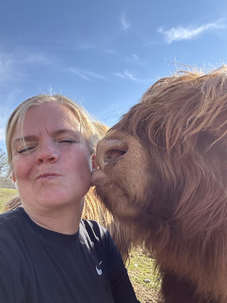 Jen, the founder of Highland Woobies, cheek-to-cheek with a red-brown Highland cow under a blue Scottish sky.